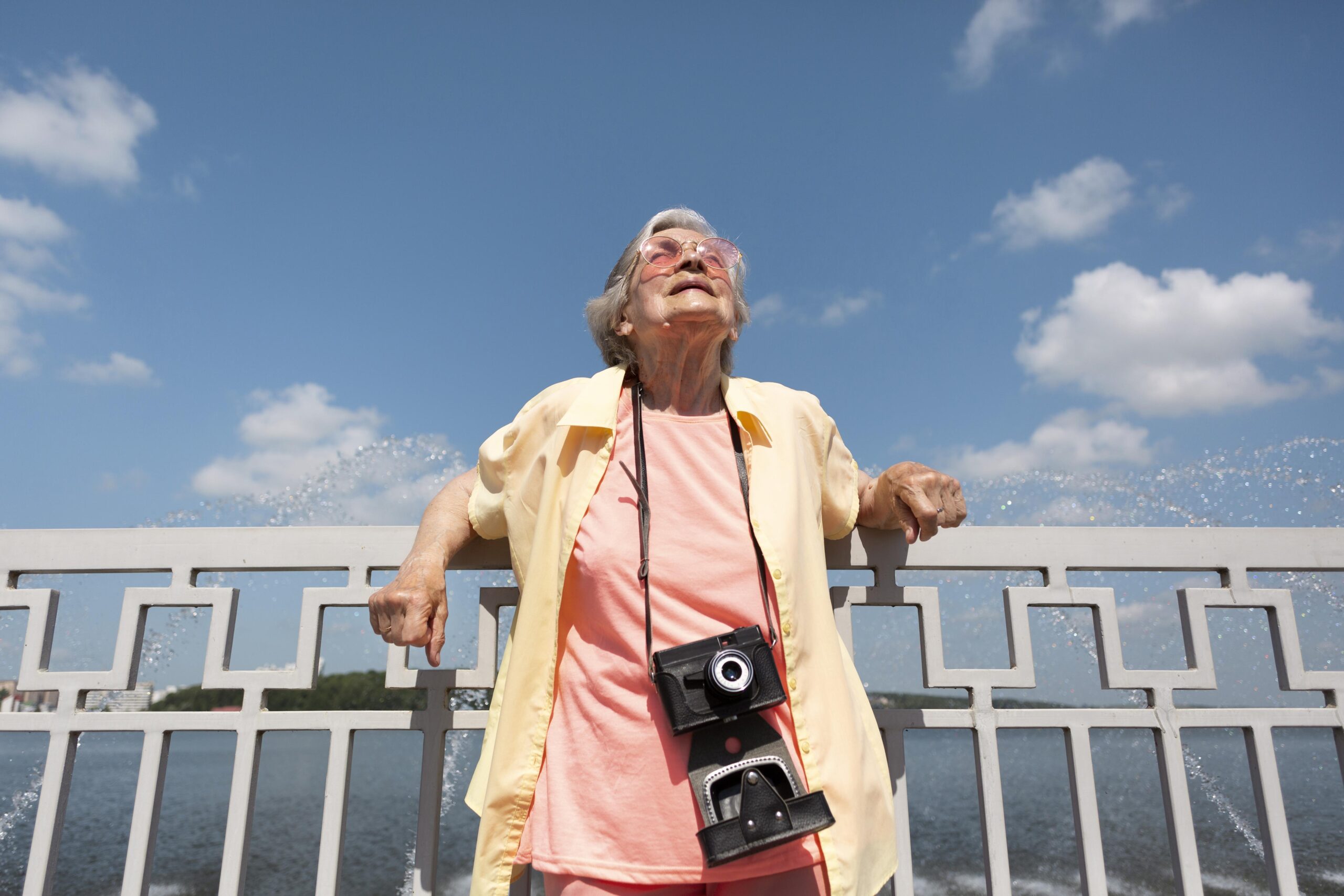 A imagem mostra uma senhora idosa com óculos escuros em local aberto olhando para cima com uma máquina fotográfica no seu pescoço